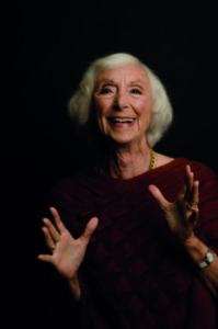 Retrato de Barbara Marx Hubbard, una mujer mayor de cabello blanco, gesticulando con ambas manos