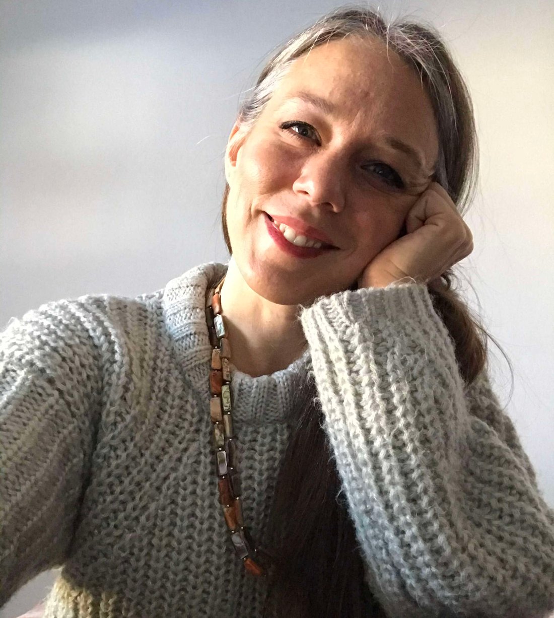 Portrait photograph of a smiling woman with grey-streaked brown hair, wearing a grey knit sweater and a beaded necklace, resting her cheek on her hand