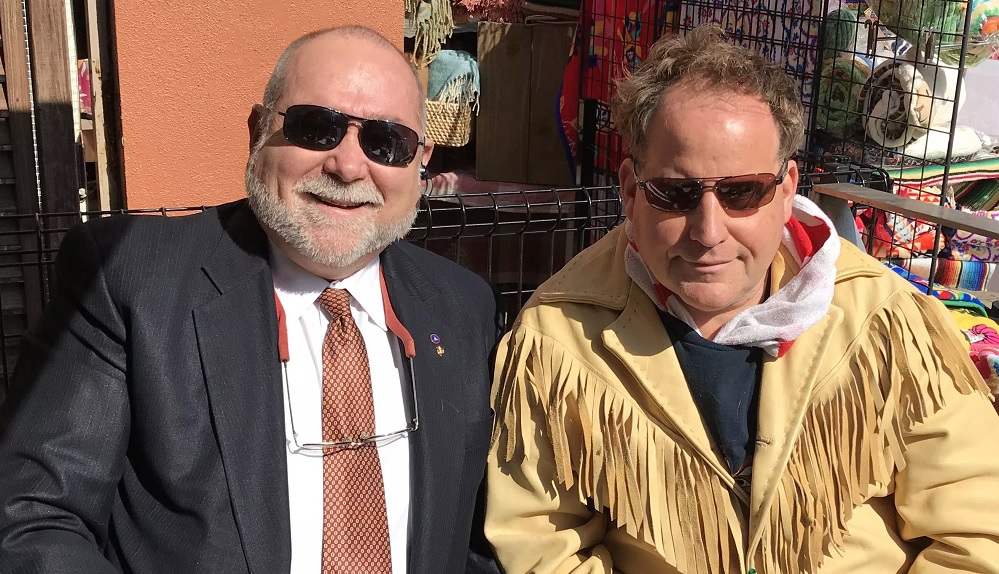 Robert David Steele con traje oscuro y lentes de sol sentado junto a Benjamin Fulford con una chamarra de gamuza con flecos, al aire libre en un puesto de mercado